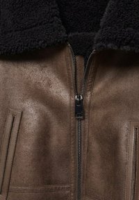 Veste en cuir marron avec col en shearling noir et fermeture éclair métallique centrale, montrant une texture détaillée et des coutures près de deux poches verticales.