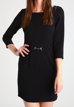 Femme portant une robe noire ajustée avec des manches trois-quarts et un petit détail de ceinture décorative à la taille.