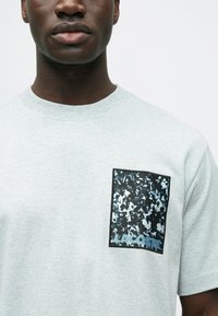 Jasnoszara koszulka z krótkim rękawem z czarną fakturowaną łatą, na której znajduje się niebieski abstrakcyjny wzór oraz napis "LACOSTE" na dole.