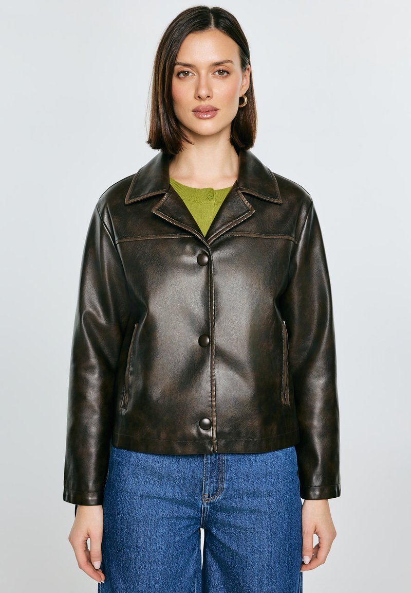 Sirgepruunkarvaline naine, kellel on kuldsed rõngaskõrvarõngad, tumepruun nahktagi rohelise topi ja siniste teksade kohal, seisab valge tausta ees.