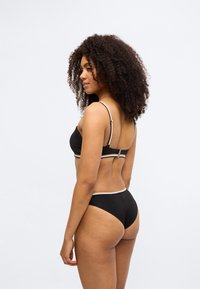 Σετ μαύρο μπικίνι με ριμπ ύφανση, λεπτές τιράντες και χρυσές διακοσμητικές λεπτομέρειες. Το πάνω μέρος έχει μια λωρίδα κάτω από το στήθος, ενώ το κάτω μέρος διαθέτει μέση μεσαίου ύψους.