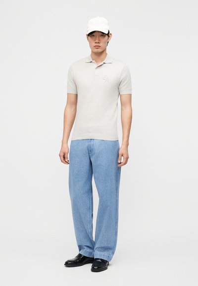 Νεαρός άνδρας με λευκό καπέλο μπέιζμπολ, ανοιχτό γκρι πόλο πουκάμισο με κοντά μανίκια, ανοιχτό μπλε τζιν και μαύρα παπούτσια, στέκεται μπροστά σε απλό φόντο.