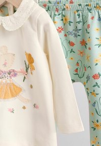 Weißes Langarmoberteil mit einem Spitzenkragen und bunten Tierstickereien, kombiniert mit hellblauen Leggings mit floralen Mustern.