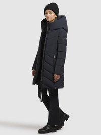 Marinblå pufferjacka med hög krage, chevronstygnmönster och dragkedja. Svarta byxor och kraftiga stövlar fulländar outfiten.
