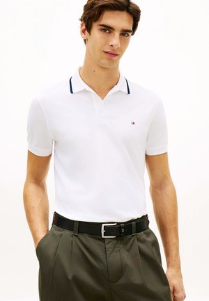 Ung mand iført en hvid poloshirt med navy krave og olivengrønne bukser med sort bælte, stående med hænderne i lommerne.