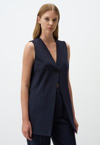 Gilet senza maniche blu navy con scollatura a V profonda, chiusura con un solo bottone e design aperto sul davanti, realizzato in un tessuto morbido con texture minimale.