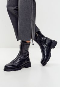 Botine din piele neagră, cu fermoare pe față și pe laterale, purtate cu pantaloni lărgi gri, pe un fundal simplu deschis.
