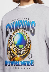 Felpa grigia con testo "CHAMPIONS" e design di un globo con un anello dorato; include "HUF WORLDWIDE" e un dettaglio per il 20° anniversario.