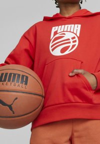 Červená mikina s kapucí a bílým logo PUMA, s přední kapsou. Držitel má basketbal s tučným černým písmem.