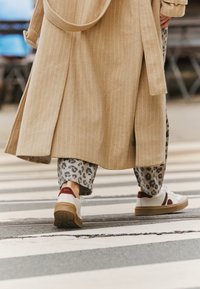 Beige striped long coat over animal print trousers and white sneakers with red and brown accents, positioned on a striped crosswalk.
