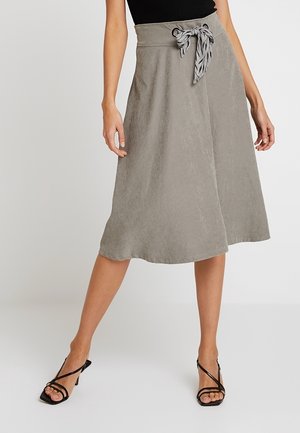 Vrouw draagt een lichtgrijze knie-lange rok met een geknoopte sjaalsriem en zwarte sandalen met hak en bandjes.
