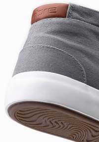 Zapato de lona gris con un parche de cuero marrón en el talón y suela de goma blanca; presenta una superficie texturizada y un patrón de tracción visible en la suela.