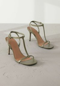 Olivengröne High Heels Sandalen mit dünnem, geflochtenem Design, quadratischer Zehenpartie und metallischem Glanz. Sie verfügen über einen schmalen Absatz und eine beigefarbene Innensohle.