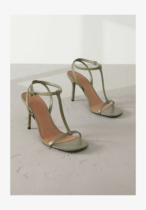 Olivengröne High Heels Sandalen mit dünnem, geflochtenem Design, quadratischer Zehenpartie und metallischem Glanz. Sie verfügen über einen schmalen Absatz und eine beigefarbene Innensohle.