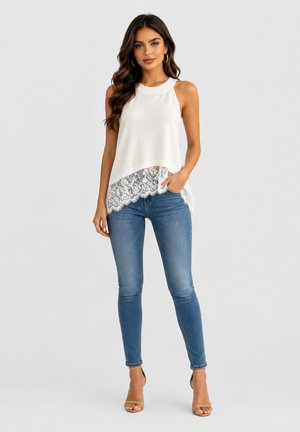 Kvinde med langt, mørkt hår iført en hvid ærmeløs top med blonderkant, blå skinny jeans og beige højhælede sandaler, stående foran en grå baggrund.
