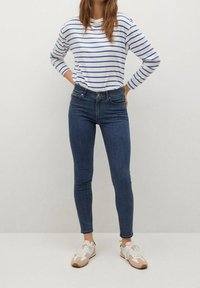 Camisa blanca de manga larga con rayas horizontales azules, combinada con jeans oscuros ajustados y zapatillas blancas con detalles en beige.