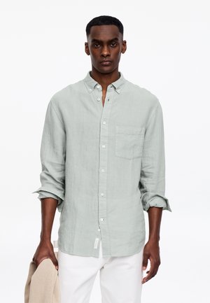 Młody mężczyzna w jasnoszarej koszuli na guziki z podwiniętymi rękawami i białych spodniach, trzymający w lewej ręce beżową kurtkę.