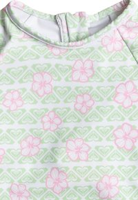 Maillot de bain à manches longues avec un motif floral rose et vert sur fond blanc, avec des coutures contrastantes et une fermeture éclair au dos.