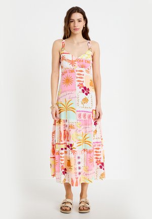 Vrouw staand, gekleed in een mouwloze, kleurrijke maxi-jurk met tropische prints en sandalen, tegen een effen witte achtergrond.