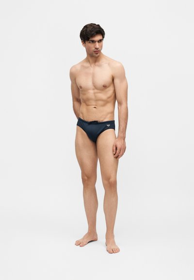 Μυώδης άνδρας στέκεται ξυπόλητος φορώντας σκούρα μπλε μαγιό με μικρό λογότυπο στο αριστερό ισχίο, μπροστά σε απλό φωτεινό φόντο.