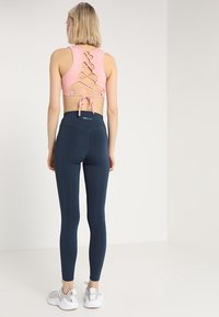 Reggiseno sportivo rosa chiaro con design a incrocio sulla schiena, abbinato a leggins blu scuro a vita alta. Il modello indossa scarpe da ginnastica bianche.