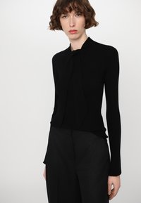 Black ribbed knit top with long sleeves and a tie detail at the neck, paired with high-waisted black trousers. Smooth texture and fitted design.
