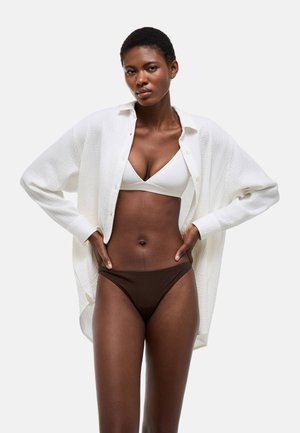 Femme portant un soutien-gorge blanc, un sous-vêtement marron et une chemise blanche texturée ouverte, debout les mains sur les hanches devant un fond uni.