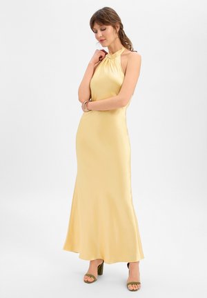 Vrouw in een mouwloze, lichtgele satijnen halterjurk met groene sandalen met hoge hakken, staand tegen een effen achtergrond.