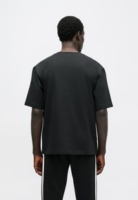 Μαύρο βαμβακερό μπλουζάκι με στρογγυλή λαιμόκοψη, κοντά μανίκια και χαλαρή εφαρμογή, σε προβολή από πίσω. Το ύφασμα έχει λεία υφή.