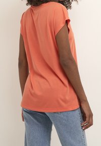 Korálové tričko s krátkým rukávem z měkké látky, s volnějším střihem a zaobleným lemem, doplněné světle modrými džínami.