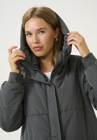 Veste grise rembourrée avec col haut et capuche. Présente des boutons-pressions et des poignets côtelés. Tissu lisse et brillant avec une finition légèrement texturée.