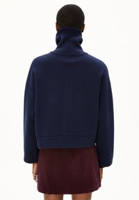 Suéter de punto grueso azul marino con cuello alto, de corte holgado y puños acanalados, usado sobre una falda burdeos. Textura suave, diseño casual.