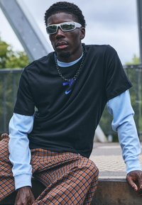 Black t-shirt layered over light blue long sleeve, checkered brown pants, sunglasses, and a chain necklace detail.