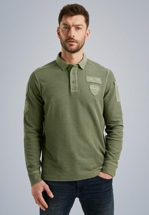 Long-sleeve olive green polo shirt with button placket, ribbed texture, shoulder patches, and embroidered logo accents. Paired with dark jeans.