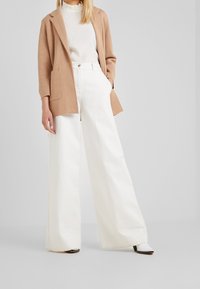 Beige Blazer über einem weißen Top, kombiniert mit weit geschnittenen weißen Hosen. Das Outfit zeichnet sich durch klare Linien und eine lockere Passform aus, mit minimalistischen Akzenten.