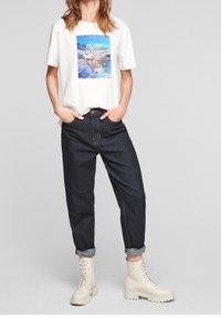 Vit t-shirt med havsgrafik och texten "VI KAN", tillsammans med mörka denimjeans uppvikta vid fållarna, samt beige snörda anklarstövlar.