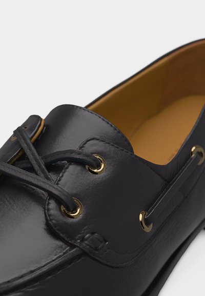 Mocassin en cuir noir avec œillets dorés et lacets noirs, doté d'une doublure intérieure couleur fauve, présenté en gros plan sur un fond blanc.