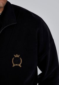 Black fleece sweatshirt with high collar and partial zip. Features gold embroidered logo with crown and laurel design, established 2012.