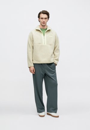 Polaire crème à demi-fermeture éclair et poche avant, portée sur une chemise verte. Pantalon ample vert foncé et baskets blanches.