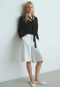 Maglione nero con un design a taglio, abbinato a pantaloni corti bianchi fino al ginocchio. L'outfit è completato da scarpe basse color crema.