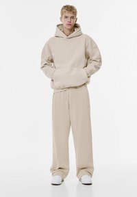 Beige Baumwollhoodie mit Kängurutasche, langen Ärmeln und verstellbarem Kapuzenzug, kombiniert mit locker sitzenden passenden Hosen. Weiße Sneakers runden den Look ab.