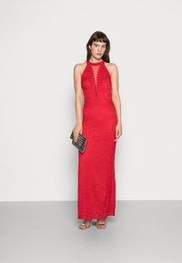 Vestido halter de renda vermelha com silhueta ajustada e detalhes transparentes. Combinado com uma carteira preta texturizada e sandálias de salto aberto prateadas.