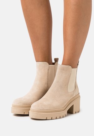 Stivaletti alla caviglia in suede beige con tacco spesso e testurizzato, pannelli laterali elastici e linguetta sul retro. Superficie liscia con una finitura opaca.