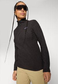 Persona che indossa un felpa nera con zip in tessuto, pantaloni beige, occhiali da sole oversize scuri, con due lunghe trecce che incorniciano il viso su uno sfondo semplice.