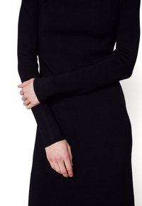 Personne portant une robe noire à manches longues, mains doucement croisées à la taille, sur fond blanc.