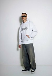 Szara bluza z kapturem z graficznym napisem, z przednią kieszenią, noszona na białej koszulce. Luźne, wyblakłe dżinsy i czarne trampki dopełniają stylizację.