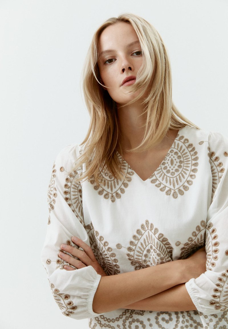 Witte linnen jurk met complexe gouden borduursels met bloem- en cirkelpatronen; V-hals ontwerp; geraffineerde mouwen met elastische manchetten.