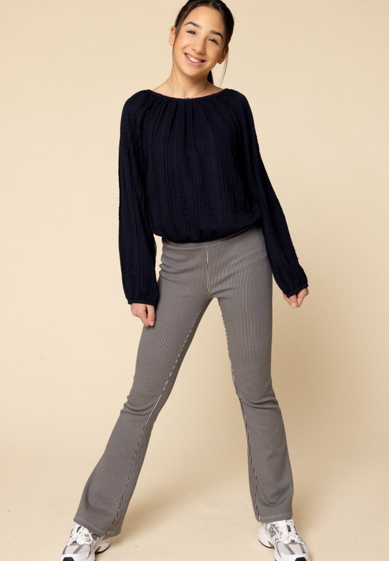 Marineblauwe gebreide lange mouwen top met een gestructureerd patroon; grijze, verticaal gestreepte flared broek. Model staat met handen op de heupen en draagt sneakers.