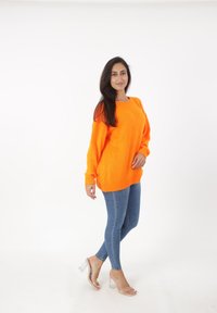 Overmaatse oranje gebreide trui met geribde manchetten en zoom, gecombineerd met slim-fit blauwe jeans en transparante hakken.