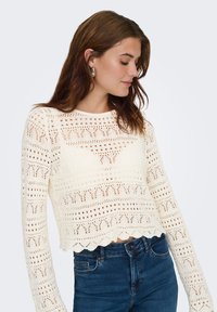 Pull crème en crochet avec manches longues et ourlet court. Présente des motifs ajourés et un bord inférieur en forme de coquille. Porté avec un jean bleu.